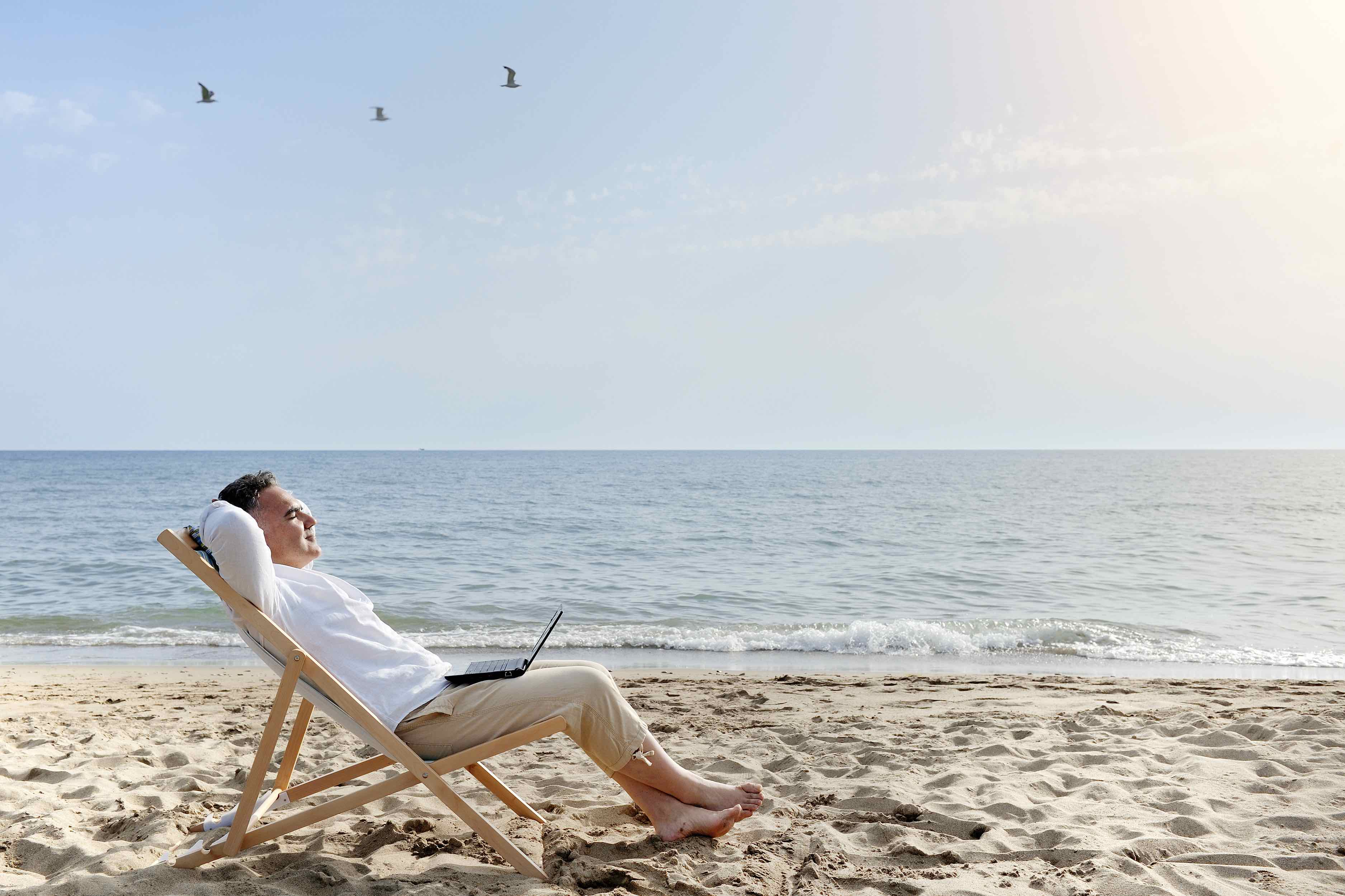 man with laptop relaxing on the beach sitting on deck chair